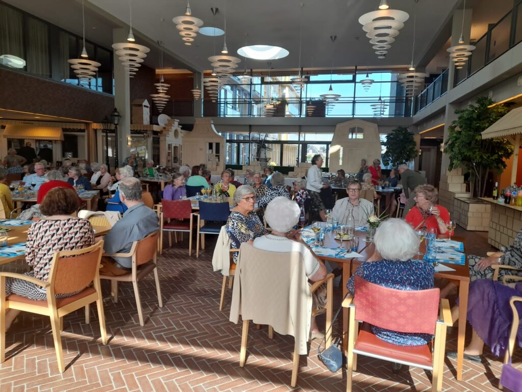 Een groep ouderen zit aan tafels in een ruime, lichte eetzaal met hoge plafonds en moderne verlichting.
