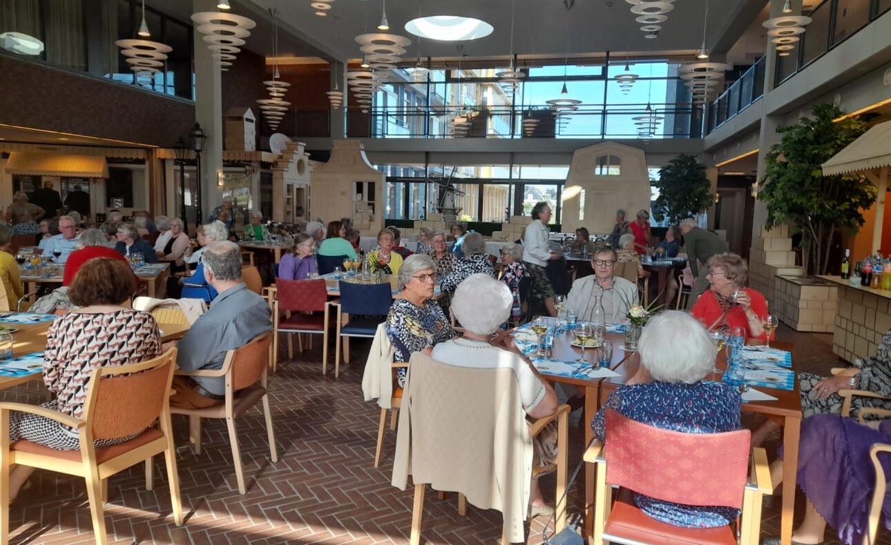 Een groep ouderen zit aan tafels in een ruime, lichte eetzaal met hoge plafonds en moderne verlichting.