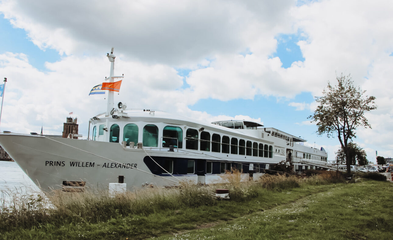 Een groot wit cruiseschip genaamd "Prins Willem-Alexander" ligt aangemeerd aan de oever, met een blauwe lucht op de achtergrond.