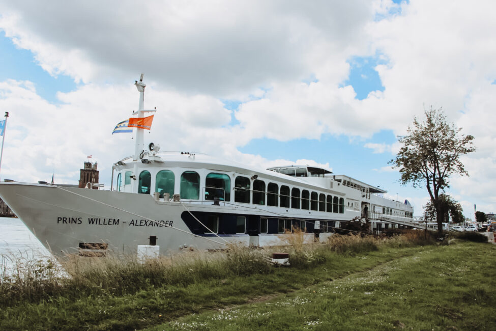 Een groot wit cruiseschip genaamd "Prins Willem-Alexander" ligt aangemeerd aan de oever, met een blauwe lucht op de achtergrond.