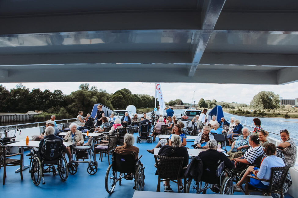 Groep mensen, waaronder ouderen in rolstoelen, geniet van een zonnige dag op een boot met uitzicht op een rivier en bomen.