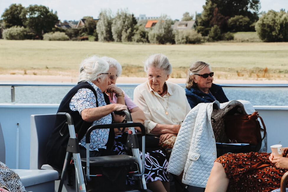 Vier oudere vrouwen zitten samen op een boot, pratend en genietend van de zonnige dag met een groen landschap op de achtergrond.