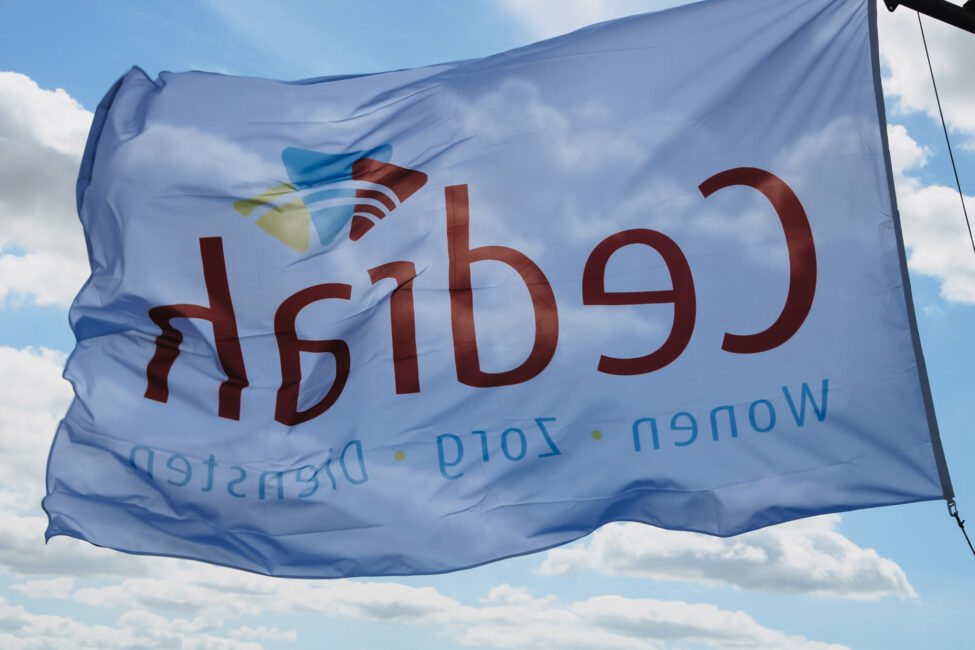 Een wapperende vlag met het logo van Cedrah tegen een blauwe lucht met wolken. De tekst op de vlag is deels leesbaar.