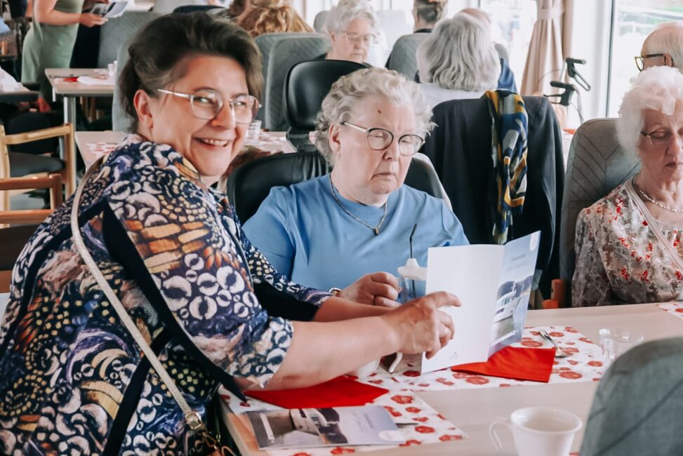 Een groep oudere vrouwen zit aan een tafel, lachend en pratend, met papieren en decoraties op de tafel voor hen.