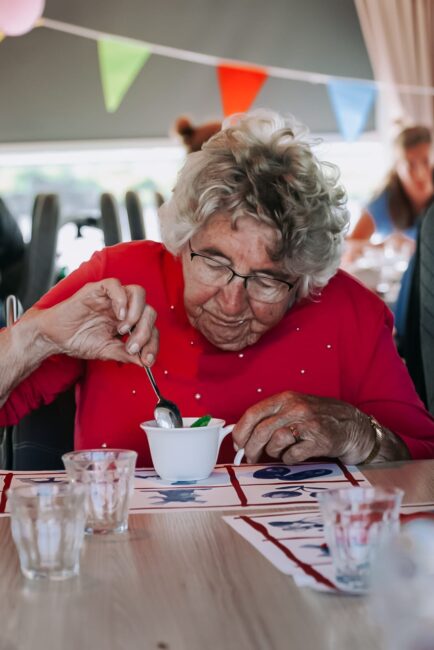 Oudere vrouw met bril en rood shirt eet dessert aan tafel, met kleurrijke vlaggetjes op de achtergrond in een feestelijke setting.