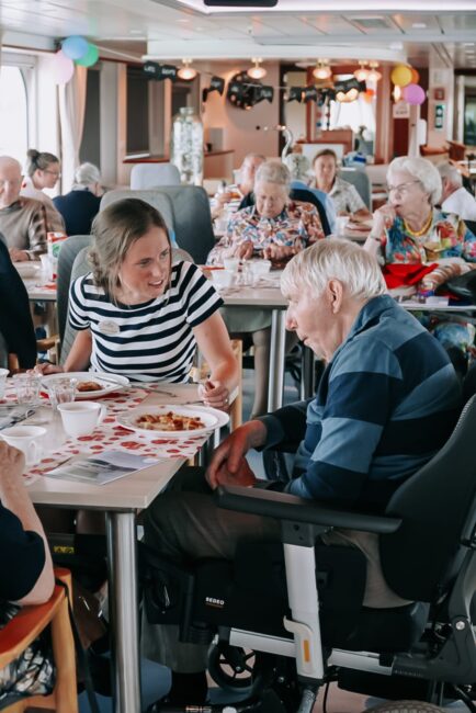 Een vrouw praat met een oudere man in een rolstoel in een drukke eetzaal. Andere ouderen zitten aan tafels op de achtergrond.