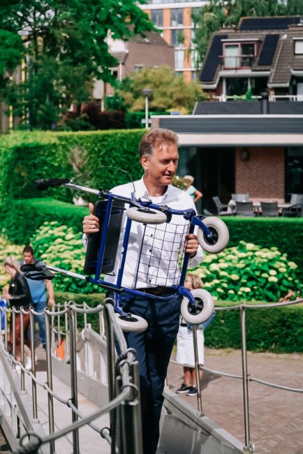 Man draagt een rollator over een loopbrug bij een groene tuin en moderne gebouwen op de achtergrond, op een zonnige dag.