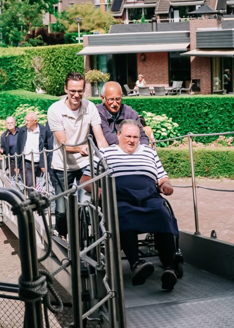 Twee mannen duwen een man in een rolstoel over een loopbrug. Op de achtergrond is een gebouw met groene struiken te zien.