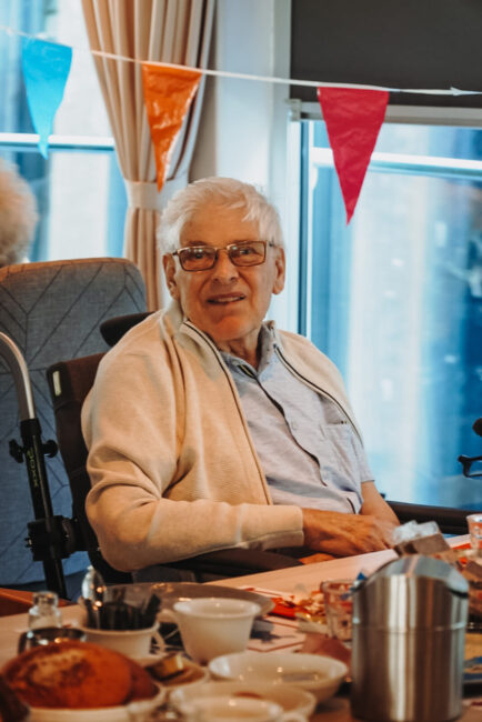 Oudere man met bril zit aan tafel met feestelijke versiering en servies, glimlachend in een gezellige omgeving.