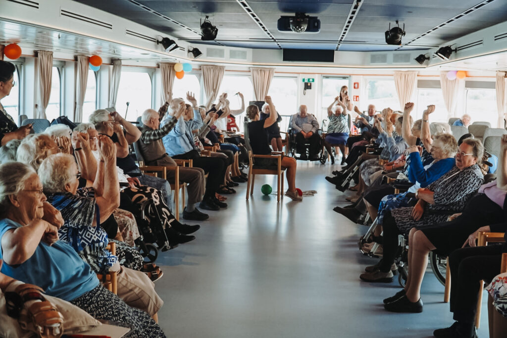 Groep ouderen doet mee aan een activiteit in een ruime zaal met ramen. Ze zitten in een kring en tillen hun armen op.