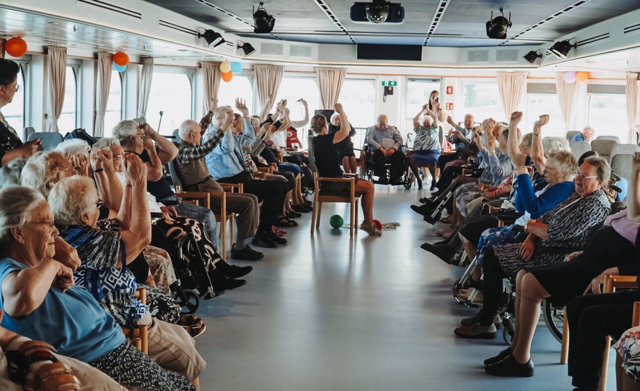 Groep ouderen doet mee aan een activiteit in een ruime zaal met ramen. Ze zitten in een kring en tillen hun armen op.