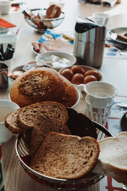 Ontbijttafel met diverse broodsoorten, eieren, en een thermoskan op een houten tafel. Servies en etenswaren zijn verspreid.