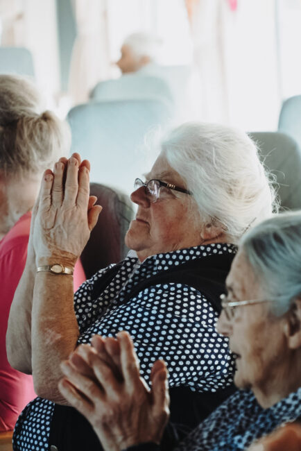 Oudere vrouw met wit haar en bril bidt met gevouwen handen, zittend naast een andere vrouw in een gemeenschappelijke ruimte.