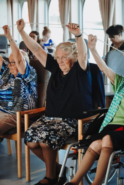 Oudere vrouwen doen samen een oefening met hun armen omhoog in een gemeenschappelijke ruimte, zittend op stoelen.