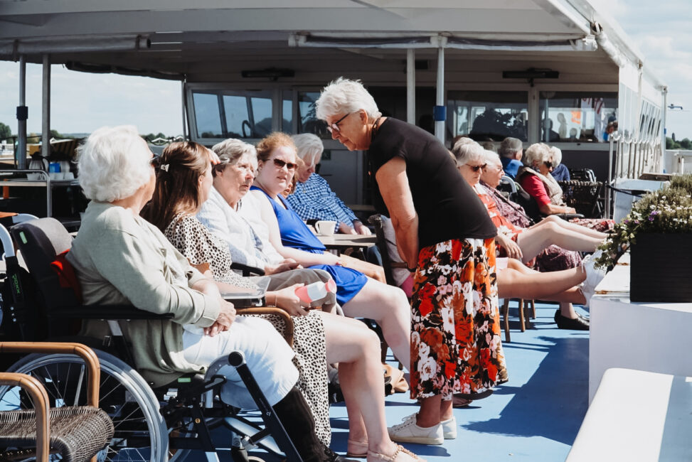 Groep ouderen geniet van een zonnige dag op een boot. Ze zitten in een rij, sommigen in rolstoelen, en praten met elkaar.