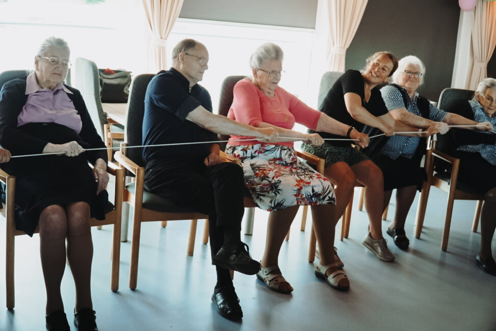 Een groep ouderen zit op stoelen in een rij en doet een activiteit met een touw, waarbij ze samen plezier lijken te hebben.