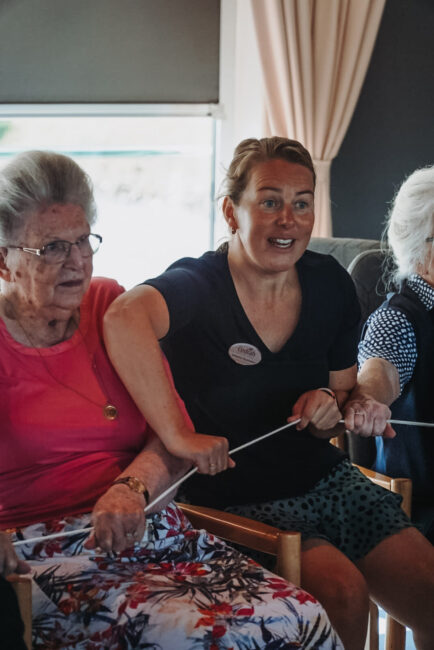 Twee oudere vrouwen en een jongere vrouw doen samen een activiteit met een touw in een kamer met gordijnen en een raam.