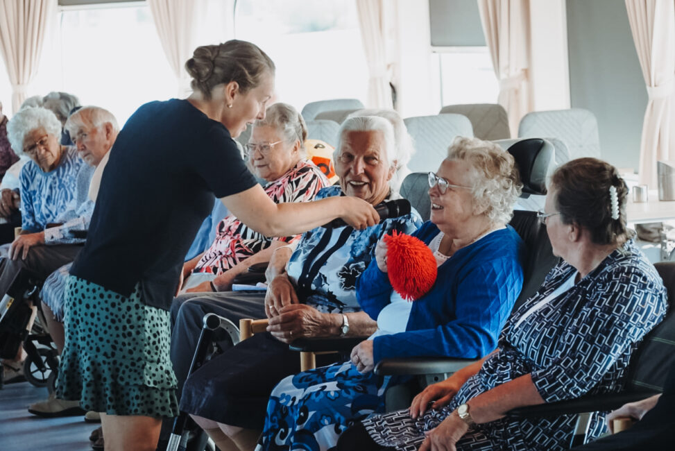 Een vrouw met microfoon interviewt een oudere vrouw in een zaal met andere senioren. Ze lachen en lijken te genieten.
