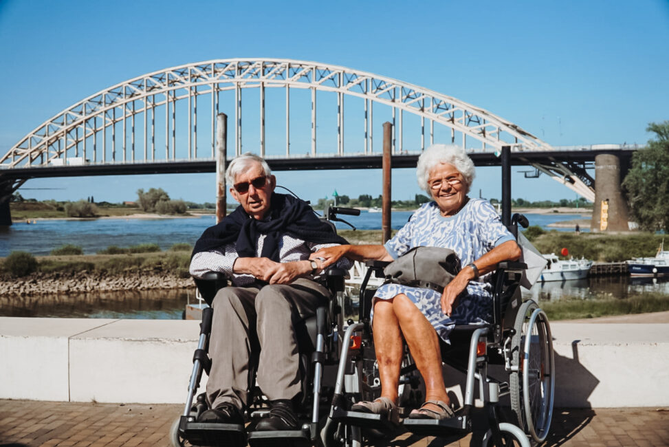 Oudere man en vrouw in rolstoelen glimlachen bij een rivier met een grote boogbrug op de achtergrond op een zonnige dag.