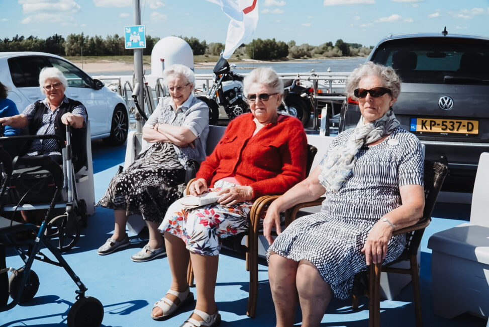 Vier oudere vrouwen zitten buiten op een zonnig terras. Ze dragen zomerse kleding en kijken ontspannen voor zich uit.