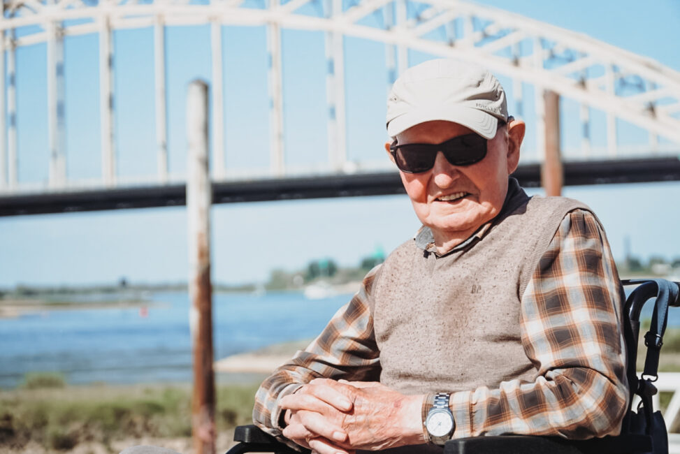Oudere man met zonnebril en pet zit in een rolstoel bij een rivier, met een brug op de achtergrond op een zonnige dag.