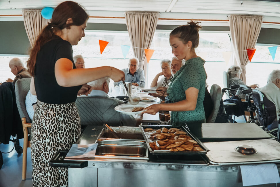 Twee vrouwen serveren eten aan een groep ouderen in een eetzaal. Op de achtergrond hangen kleurrijke vlaggetjes.