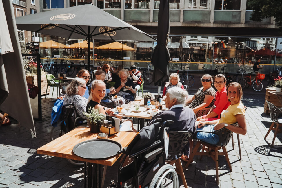 Groep mensen zit buiten op een terras aan een tafel, genietend van eten en drinken op een zonnige dag in de stad.