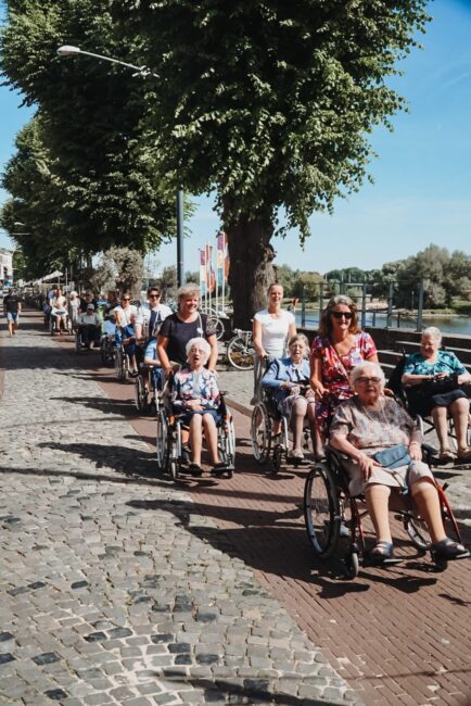 Groep mensen in rolstoelen met begeleiders wandelen over een geplaveide straat langs een rivier op een zonnige dag.