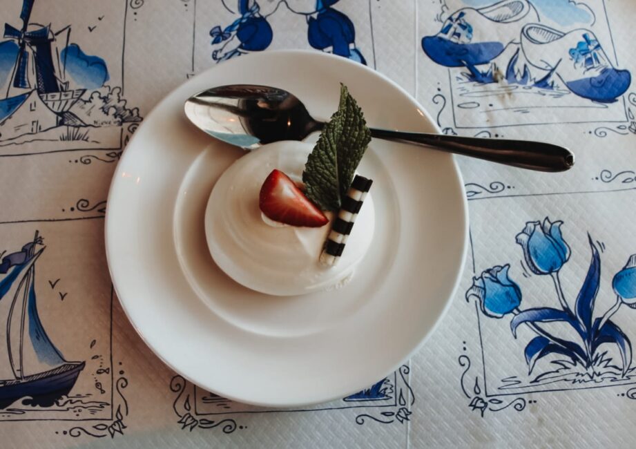Een dessert met room, een halve aardbei, een chocoladestokje en muntblaadje op een wit bord met een lepel ernaast.