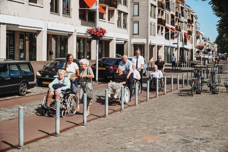 Groep mensen duwt rolstoelen op een zonnige dag in een stad. Op de achtergrond zijn gebouwen en geparkeerde fietsen te zien.