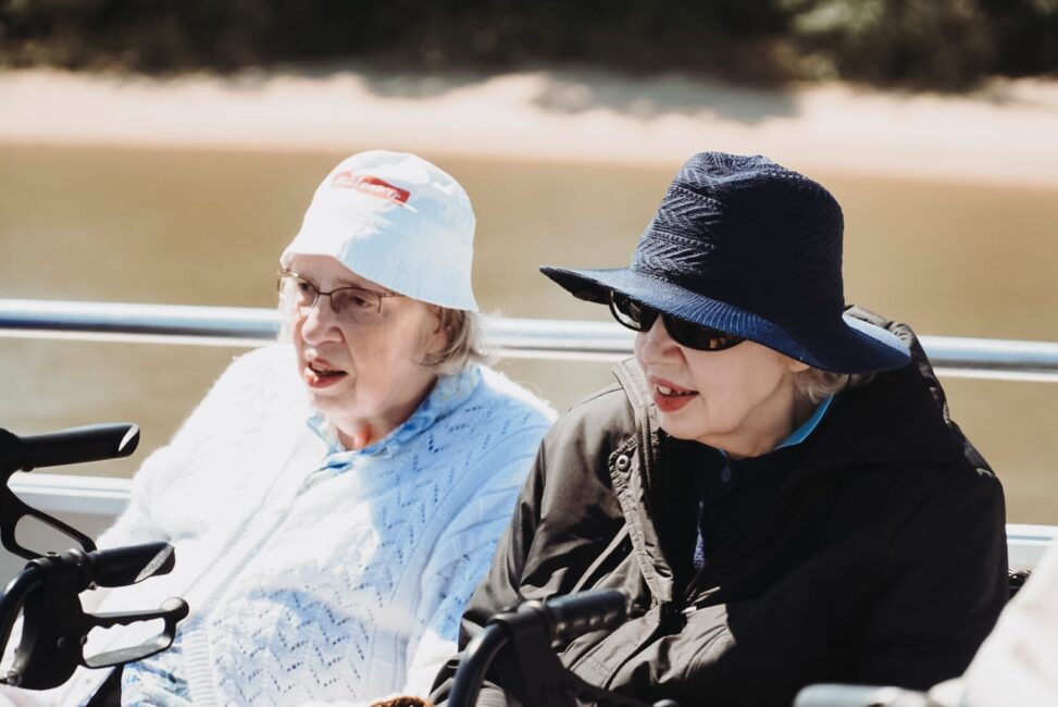 Twee oudere vrouwen met hoeden zitten buiten op een zonnige dag, genietend van een boottocht met een rustige rivier op de achtergrond.