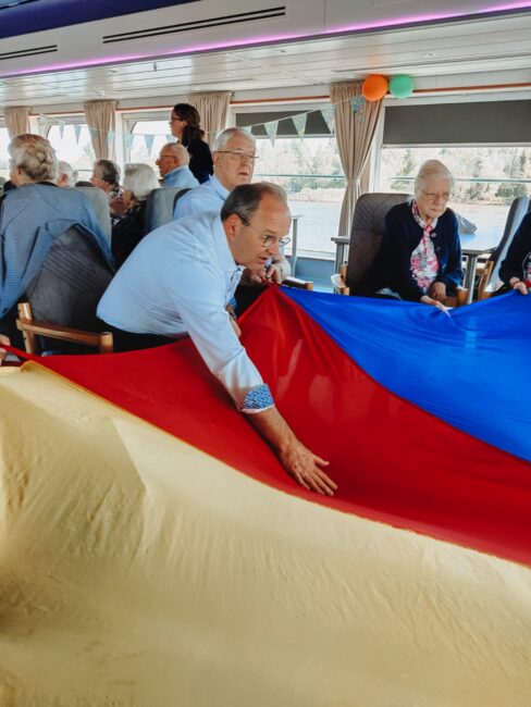 Een groep ouderen zit in een kamer terwijl een man een grote, kleurrijke parachute vasthoudt. De sfeer is ontspannen en vrolijk.