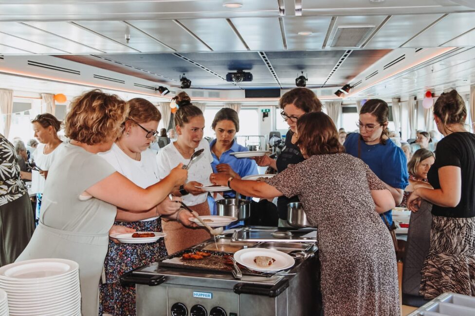 Groep mensen verzameld rond een buffet op een boot, terwijl ze eten opscheppen. De sfeer is ontspannen en gezellig.