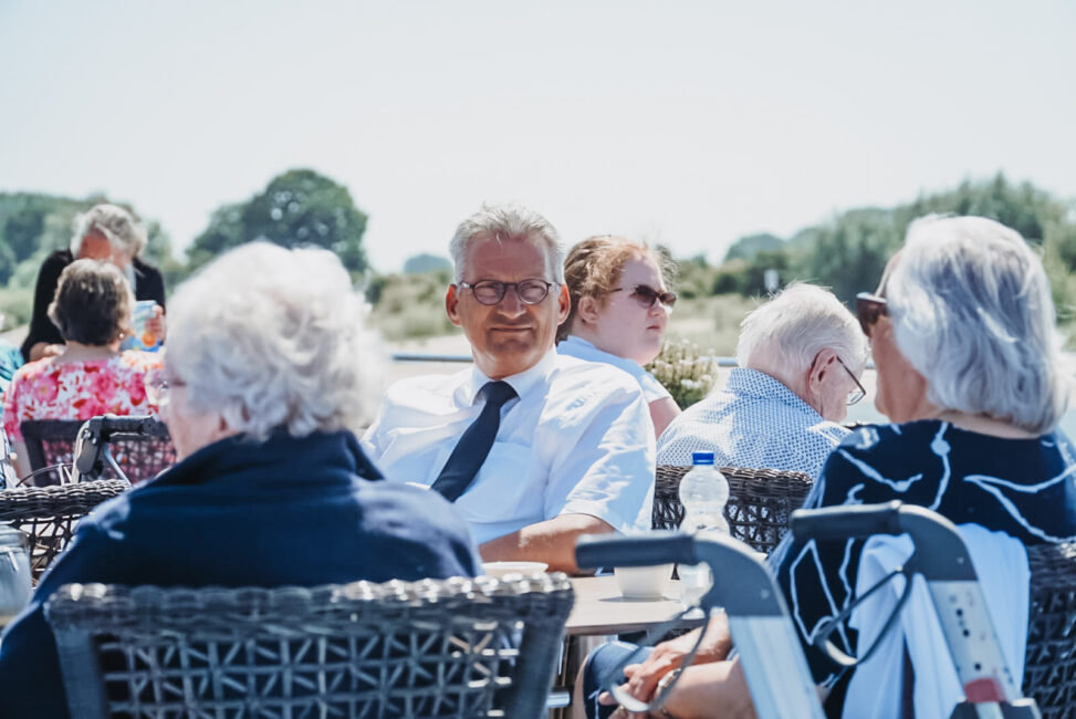 Groep mensen zit buiten op een terras. Een man in een pak en een vrouw met zonnebril zijn in gesprek. Zonnige dag.
