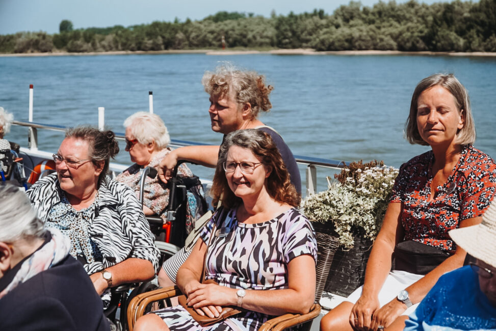 Groep oudere vrouwen geniet van een zonnige dag op een boot, met een rivier en groene bomen op de achtergrond.