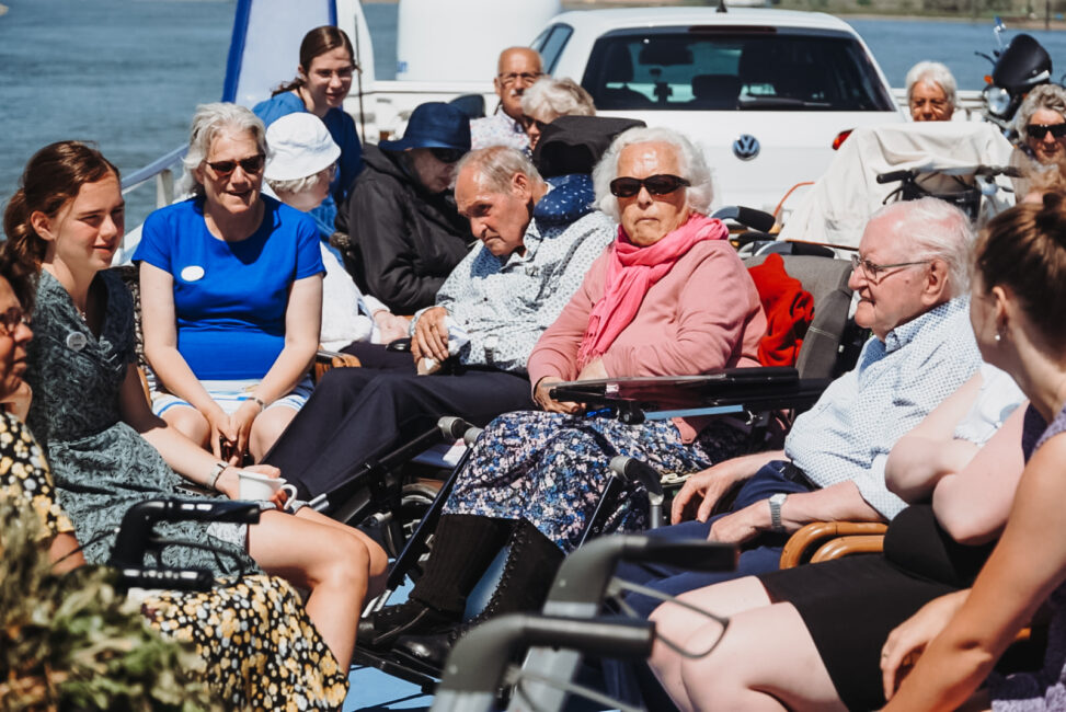 Groep ouderen en begeleiders genieten van een zonnige dag op een boot. Ze zitten samen en kijken ontspannen om zich heen.