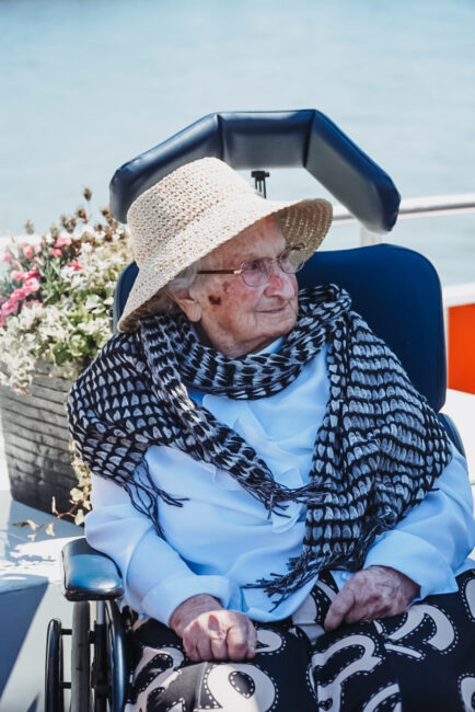 Oudere vrouw in een rolstoel met een hoed en sjaal, genietend van een zonnige dag op een boot, met bloemen op de achtergrond.