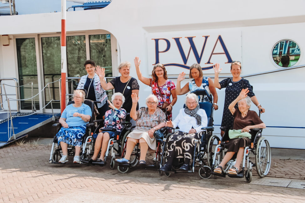Groep ouderen en verzorgers poseert lachend en zwaaiend voor een boot met de letters "PWA" op de zijkant.