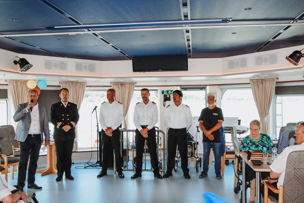Een groep mensen staat op een podium in een zaal met publiek. Enkele personen dragen uniformen. Tafels en stoelen zijn zichtbaar.