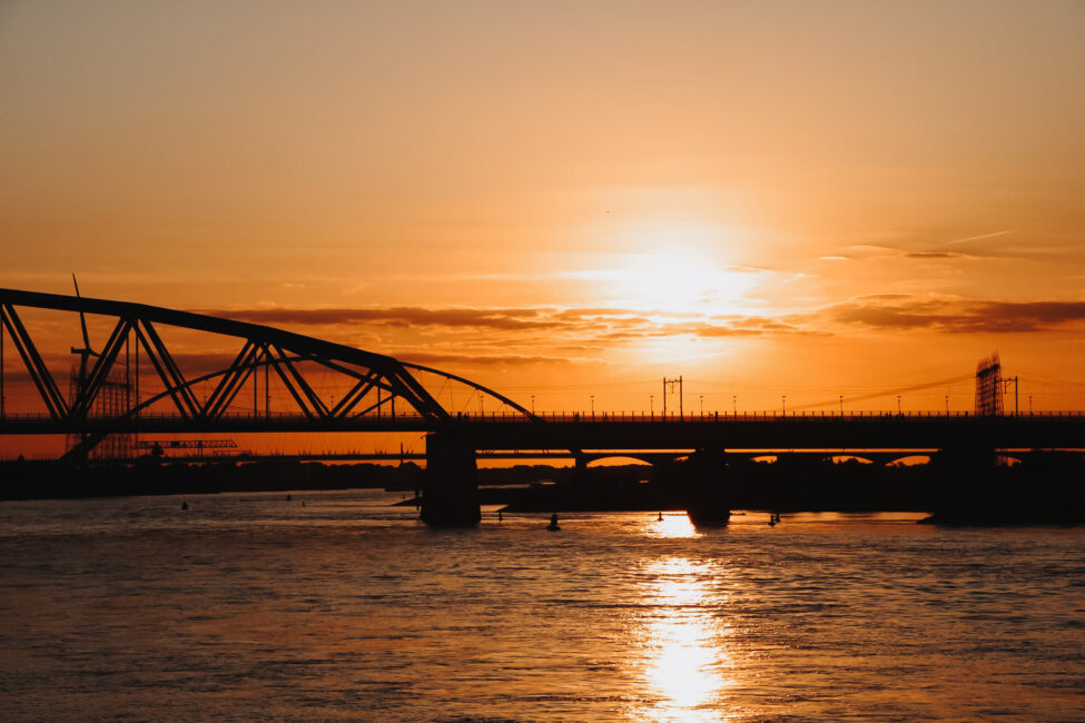 Zonsondergang boven een rivier met een silhouet van een brug. De lucht is oranje en weerspiegelt op het wateroppervlak.