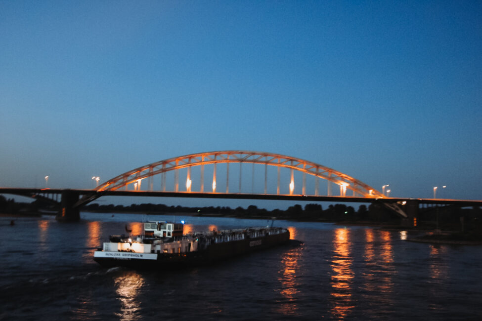Een verlichte brug bij schemering met een vrachtschip dat over de rivier vaart, weerspiegelingen van licht op het water.