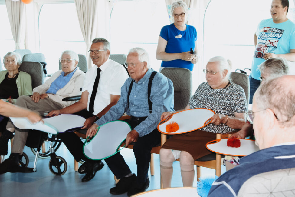 Groep ouderen speelt een spel met kleurrijke ballen en ronde schijven in een lichte ruimte. Iedereen lijkt betrokken en vrolijk.