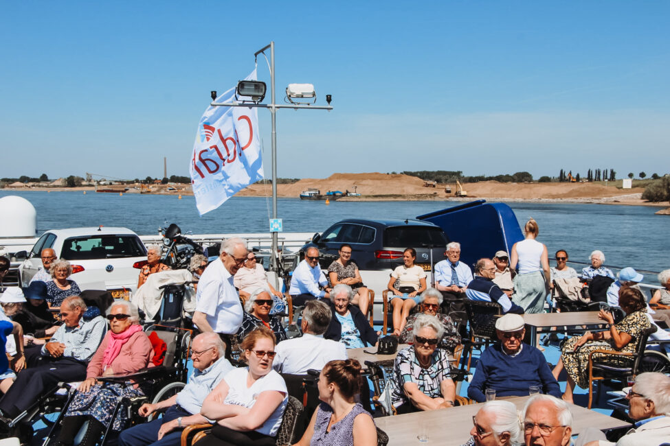Groep mensen geniet van een zonnige dag op een veerboot met een Cedrah-vlag, varend over een brede rivier onder een blauwe hemel.
