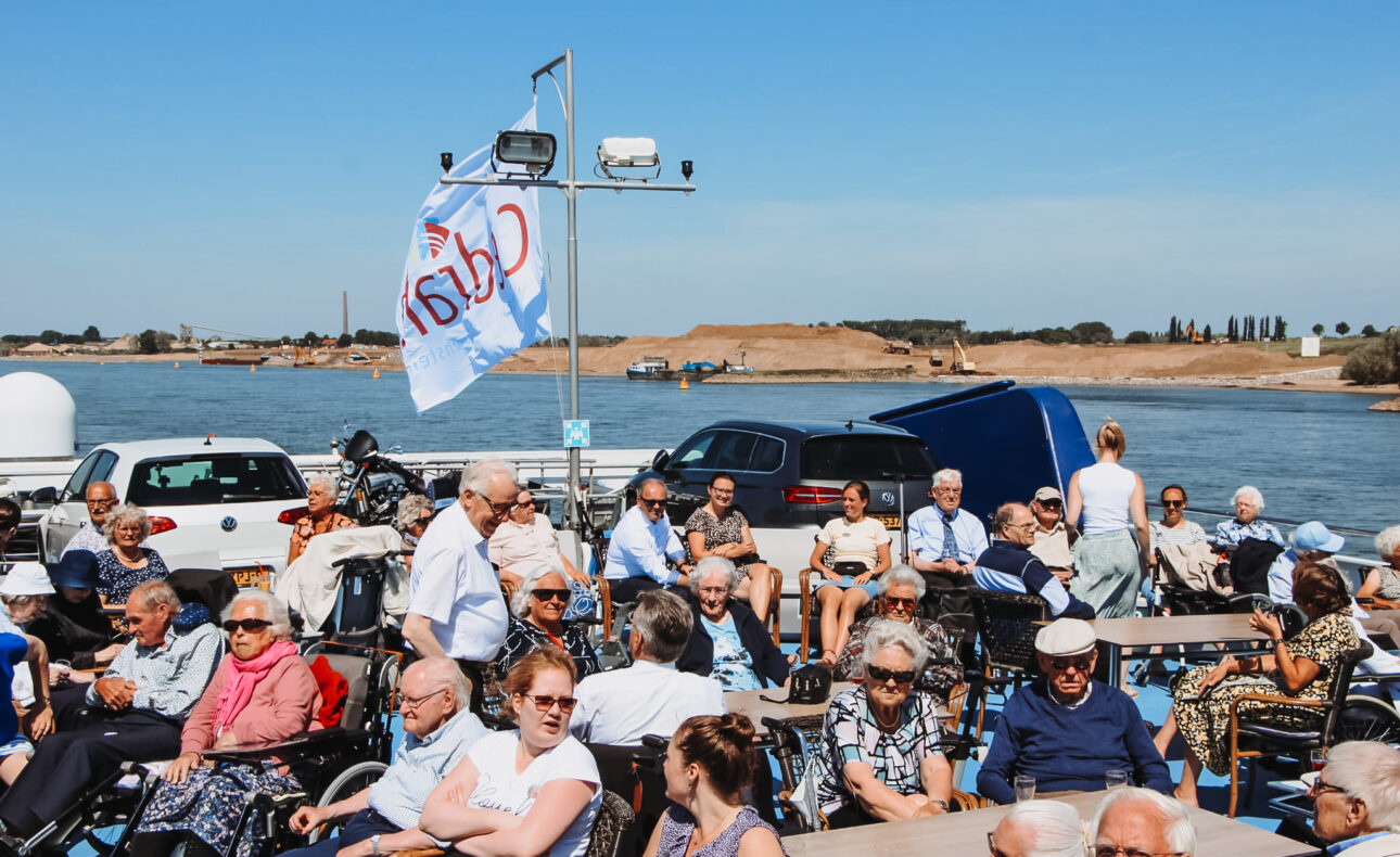 Groep mensen geniet van een zonnige dag op een veerboot met een Cedrah-vlag, varend over een brede rivier onder een blauwe hemel.