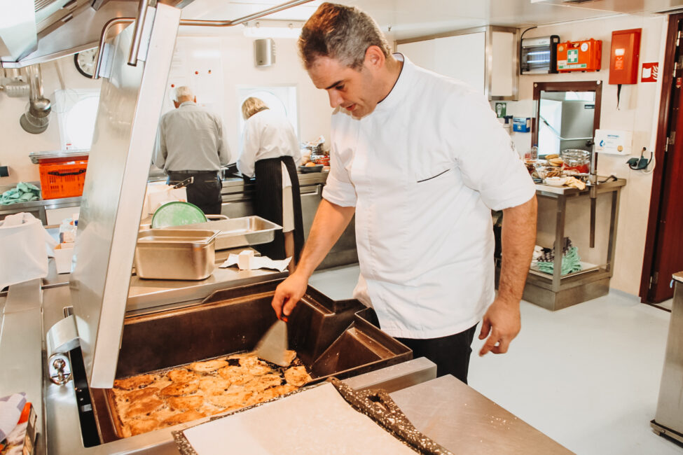 Een kok in een witte jas bereidt een gerecht in een grote pan in een professionele keuken. Andere koks werken op de achtergrond.