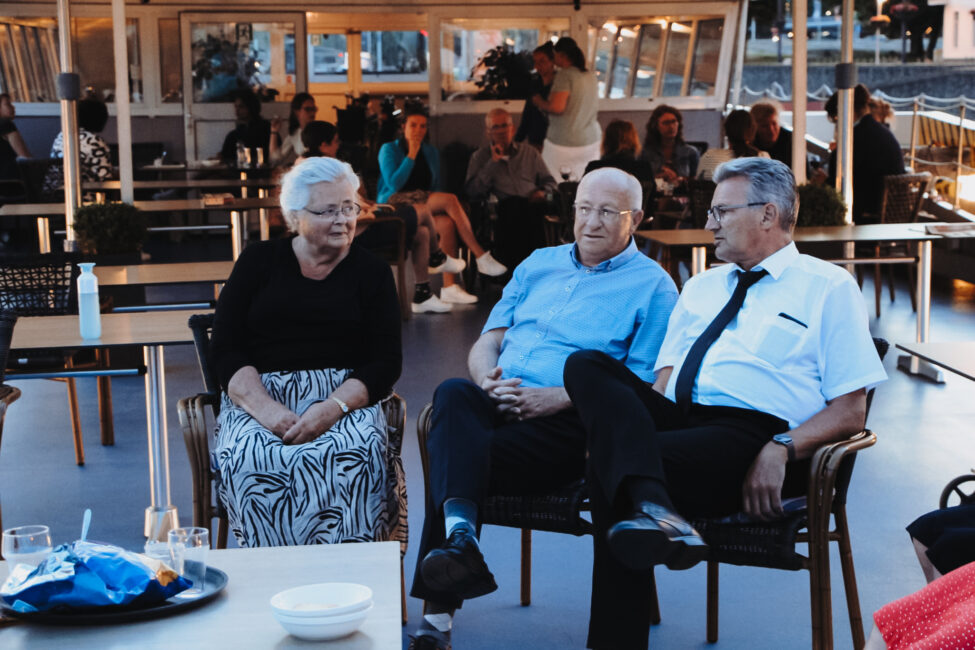 Drie oudere mensen zitten samen op een boot, gezellig pratend. Op de achtergrond zijn andere mensen en tafels zichtbaar.