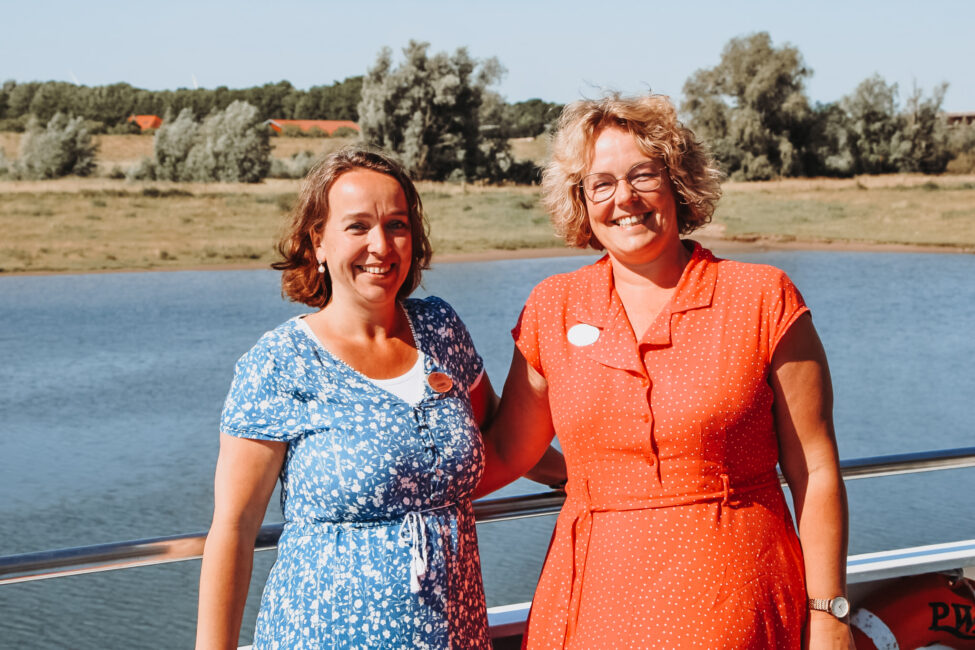 Twee vrouwen glimlachen naar de camera terwijl ze buiten op een zonnige dag voor een meer en bomen op de achtergrond staan.