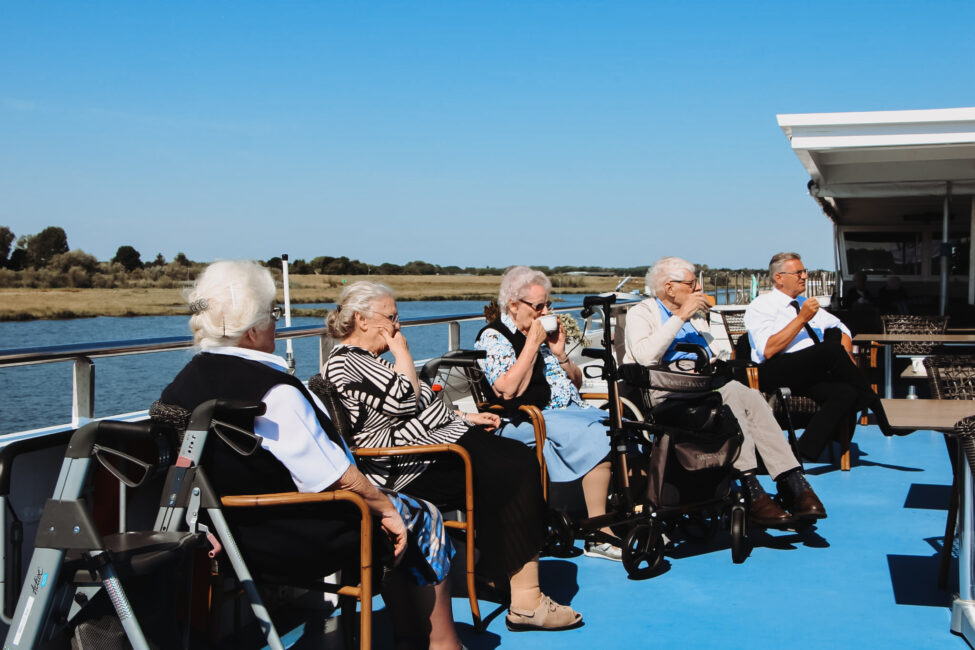Groep ouderen geniet van een zonnige dag op een boot, zittend in stoelen met uitzicht op het water en de groene oever.