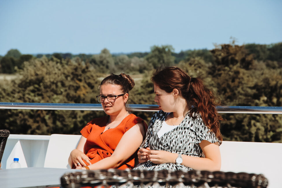 Twee vrouwen zitten buiten op een bank met bomen op de achtergrond. De vrouw links draagt een oranje jurk, de vrouw rechts een grijze.