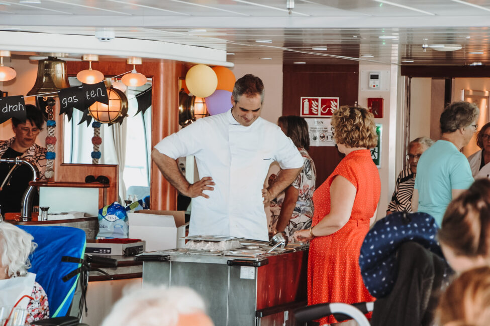 Een kok in een witte jas praat met een vrouw in een rode jurk tijdens een evenement in een drukke ruimte met mensen.